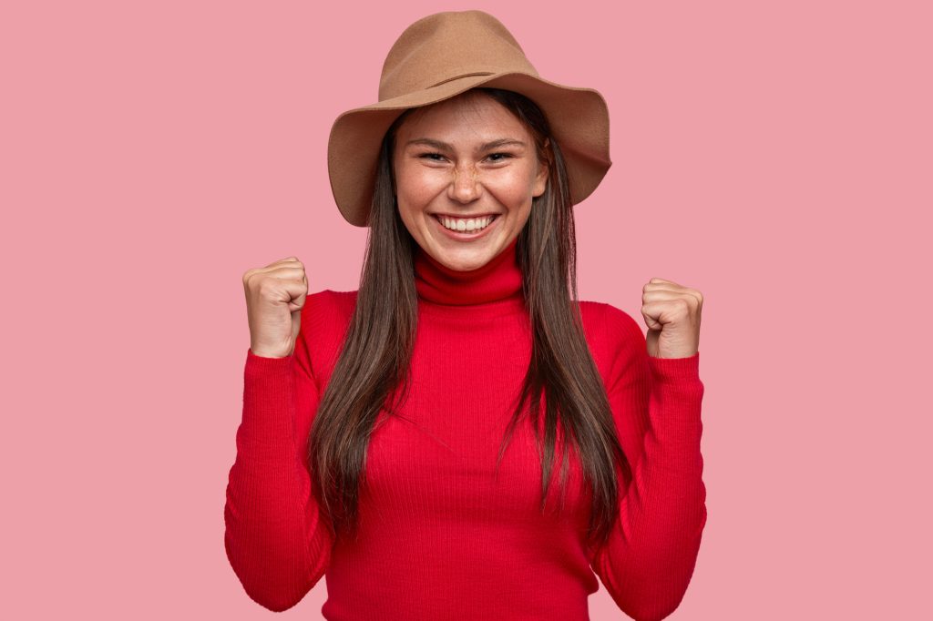 Cheerful freckled Caucasian woman raises hands clenched in fists, has toothy smile, rejoices positiv