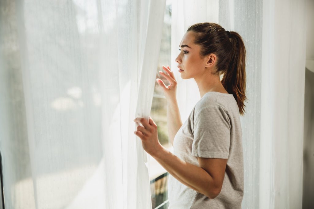 Woman Feeling Anxious While Standing Near The Window At Home